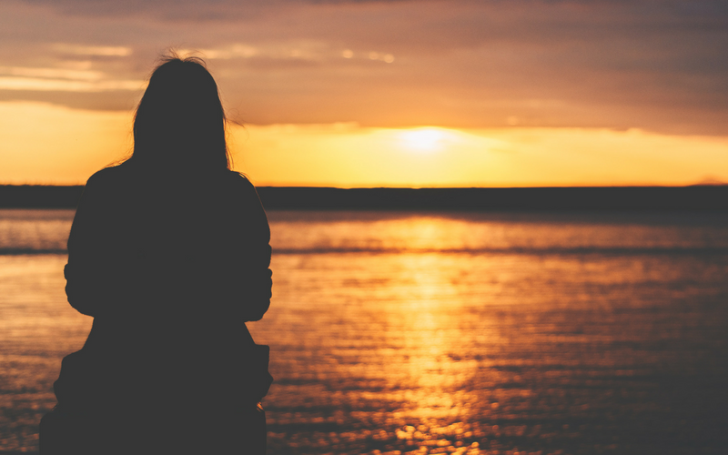 The outline of a woman is in the foreground as an orange sunset is reflected on the ocean.