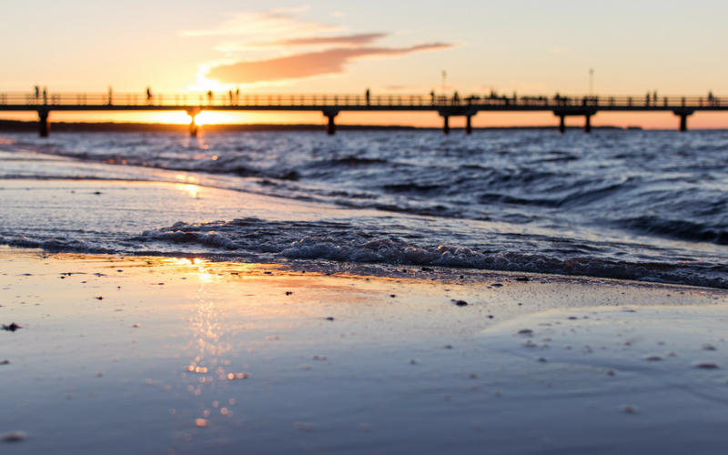 The sun sets behind a pier as the ocean tides roll in on the beach.