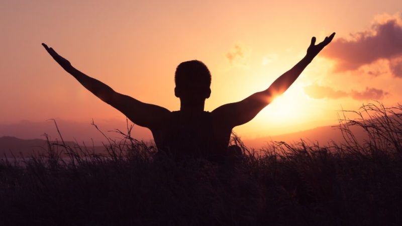 man looking out at the sun with arms up